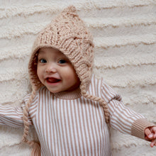 Load image into Gallery viewer, baby smiling laying on white blanket wearing latte colored baby knit bonnet and white and tan striped long sleeved onesie