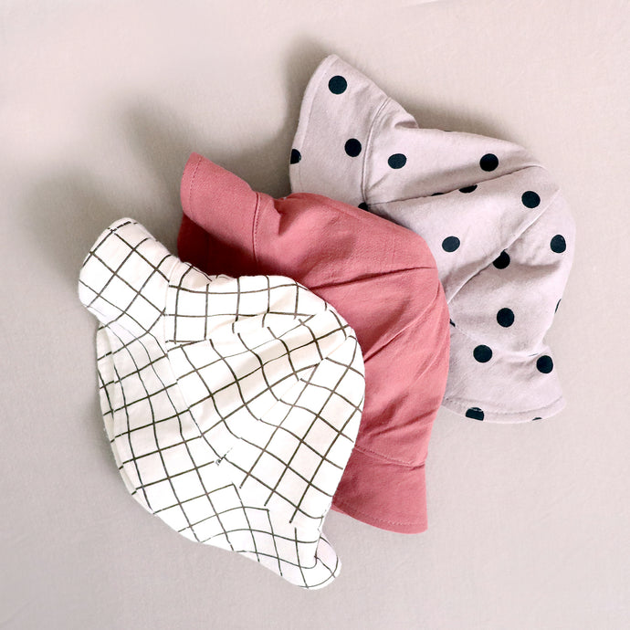 Three baby cotton bucket hats. Brown and white grid pattern, solid red, and beige with black polka dots. 