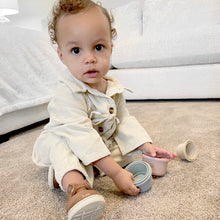 Load image into Gallery viewer, baby sitting on carpeted floor in front of white couch looking at camera playing with cups wearing white corduroy jumpsuit and brown boots 

