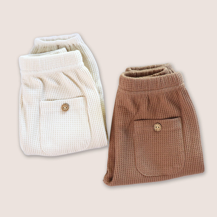 Cream and brown baby waffle pants folded next to each other showing back pocket with button