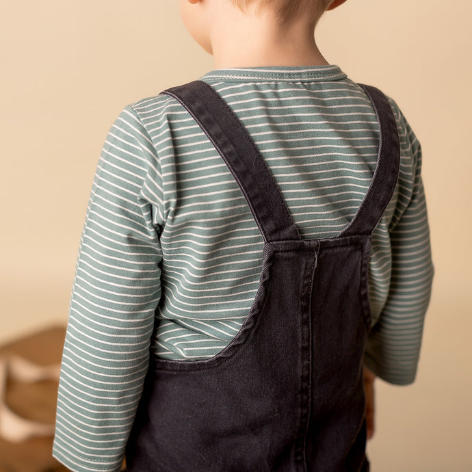 toddler standing wearing green striped long sleeved shirt and dark denim overalls