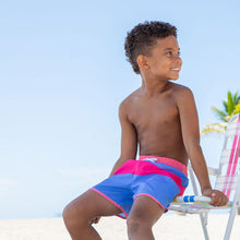 Load image into Gallery viewer, child sitting on beach wearing pink and blue striped swim trunks
