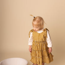 Load image into Gallery viewer, toddler standing looking down wearing white long sleeved shirt and yellow gold gingham sleeveless tiered dress

