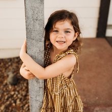 Load image into Gallery viewer, child hugging a tree wearing gold gingham tiered toddler dress with hand tied bow straps
