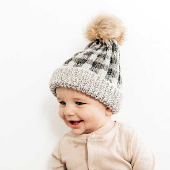 Baby smiling wearing grey buffalo checkered knit hat with ribbed cuff and fur pom pom topper and beige knit top
